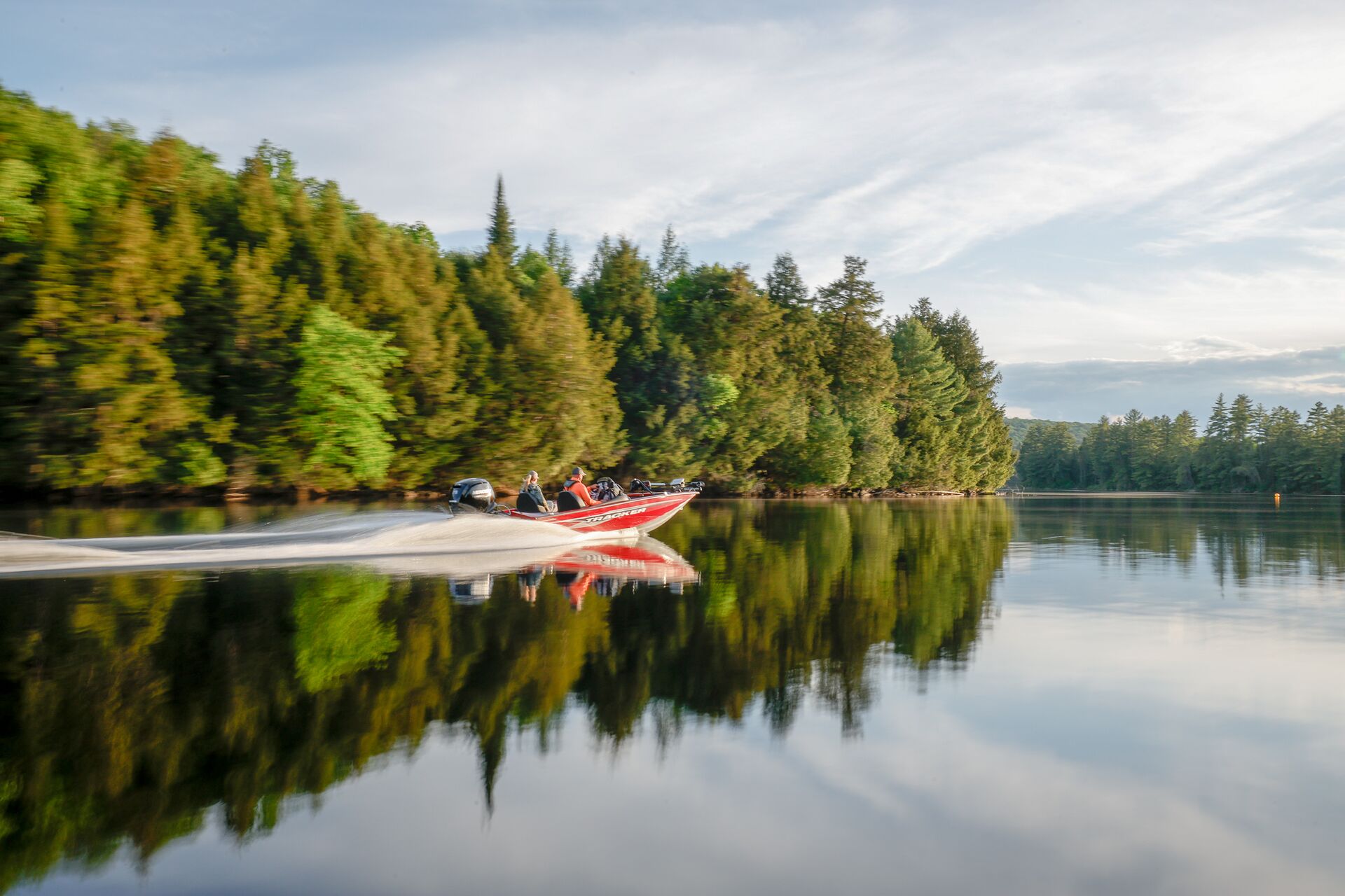 Red fishing boat moves along treelined lake shore.