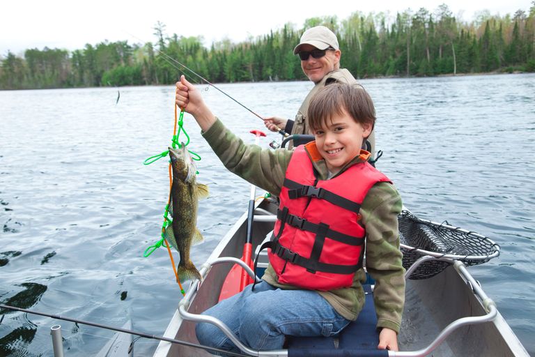 A child with an adult in a boat holds up a walleye fish.